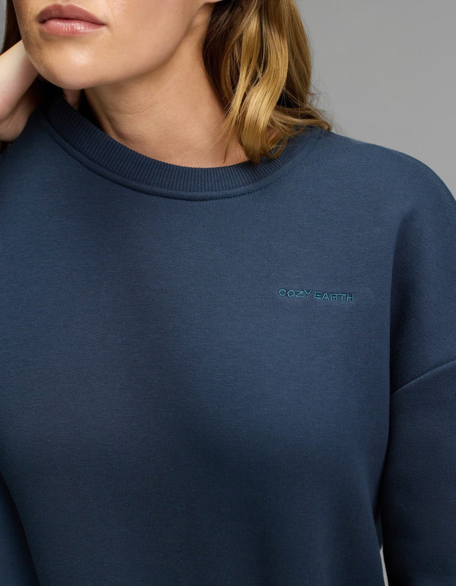 A person wearing the Cozy Earth Women’s CityScape Crewneck in dark blue, featuring small, light blue “Cozy Earth” embroidery on the chest. Only their lower face, neck, and upper torso are visible.