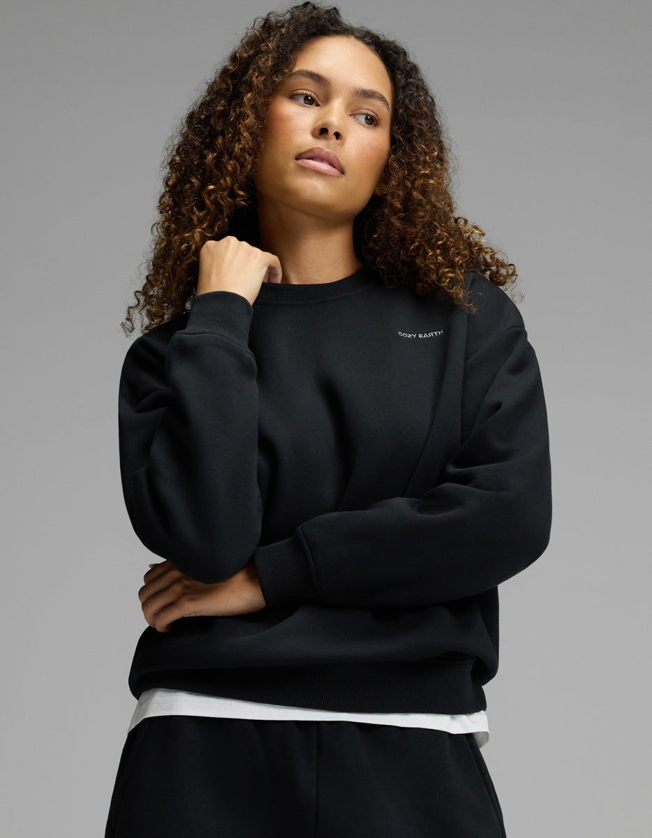 Wearing the Cozy Earth Women’s CityScape Crewneck in black over a white shirt, a woman with curly hair stands against a plain gray background, her right hand touching her chin and her left arm crossed over her chest.|Color:Jet Black