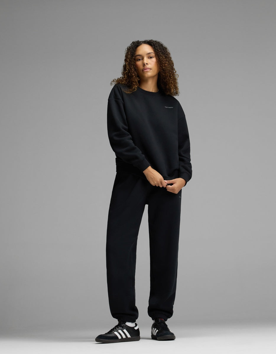 A person with curly hair stands against a plain gray background, wearing the Cozy Earth Women’s CityScape Crewneck in black, matching black sweatpants, and black Adidas sneakers with white stripes.