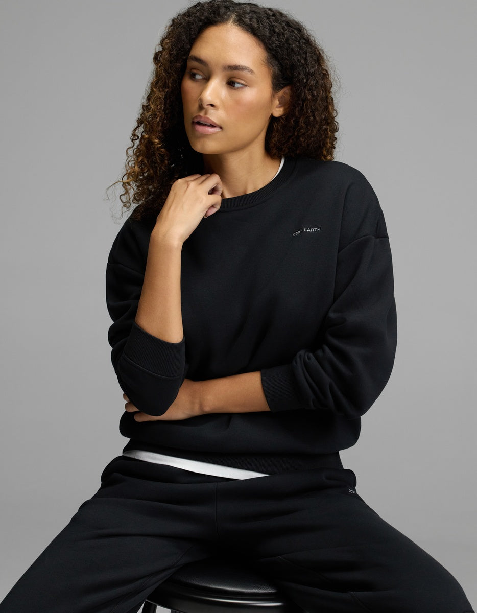 A woman with curly hair wears the Cozy Earth Women’s CityScape Crewneck and matching pants, sitting on a stool against a gray background, gazing to the side with one hand resting on her chin.