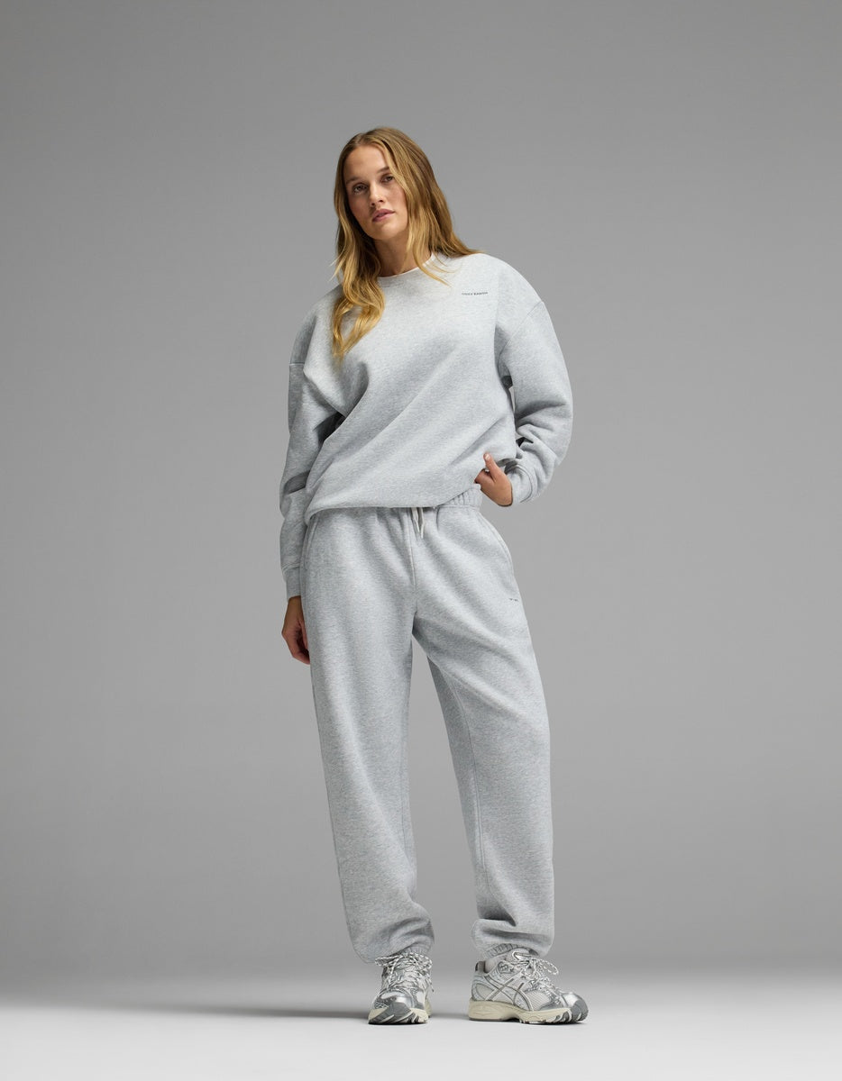 A woman stands against a plain gray background wearing the Cozy Earth Women’s CityScape Crewneck & Sweatpant Set and silver athletic sneakers, with one hand in her pocket.