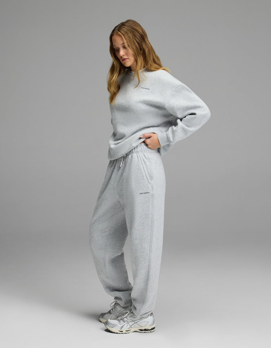 A woman stands in profile wearing the Cozy Earth Women’s CityScape Crewneck & Sweatpant Set in light gray, paired with gray sneakers. She has long, light brown hair and looks down with one hand in her pocket against a plain gray background.