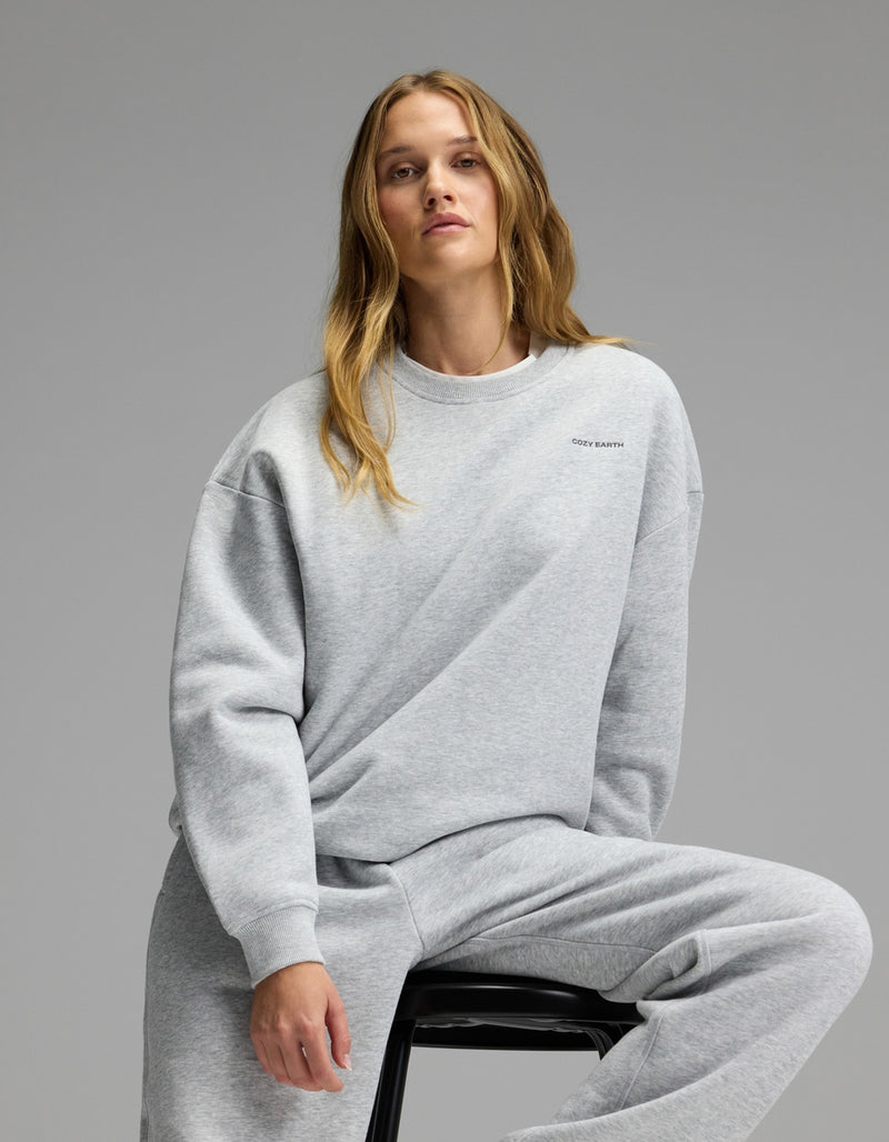 A woman with long blonde hair sits on a stool, wearing the Cozy Earth Women’s CityScape Crewneck & Sweatpant Set in light gray, against a plain gray background.