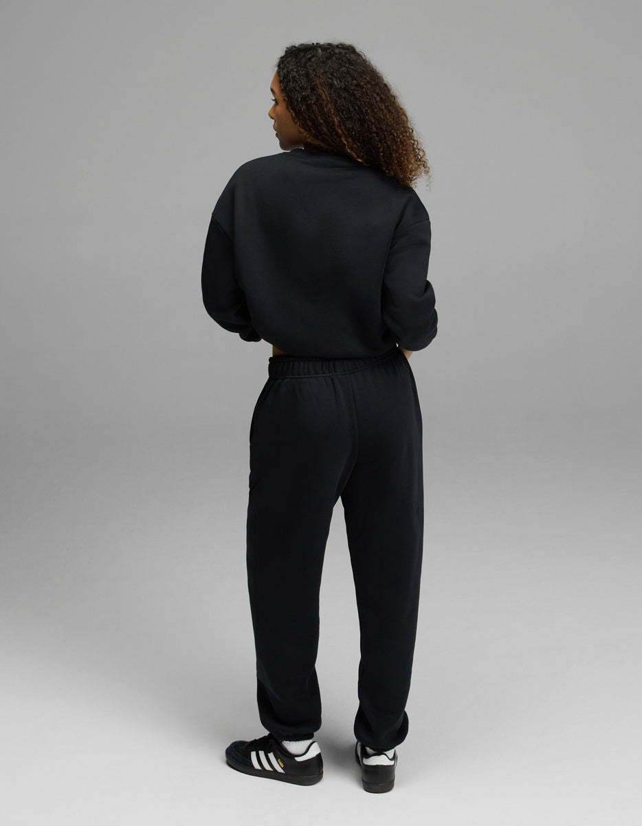 A person with curly hair, seen from the back, is wearing the Women’s CityScape Crewneck & Sweatpant Set by Cozy Earth in black, along with black Adidas sneakers featuring white stripes, standing on a light gray background.