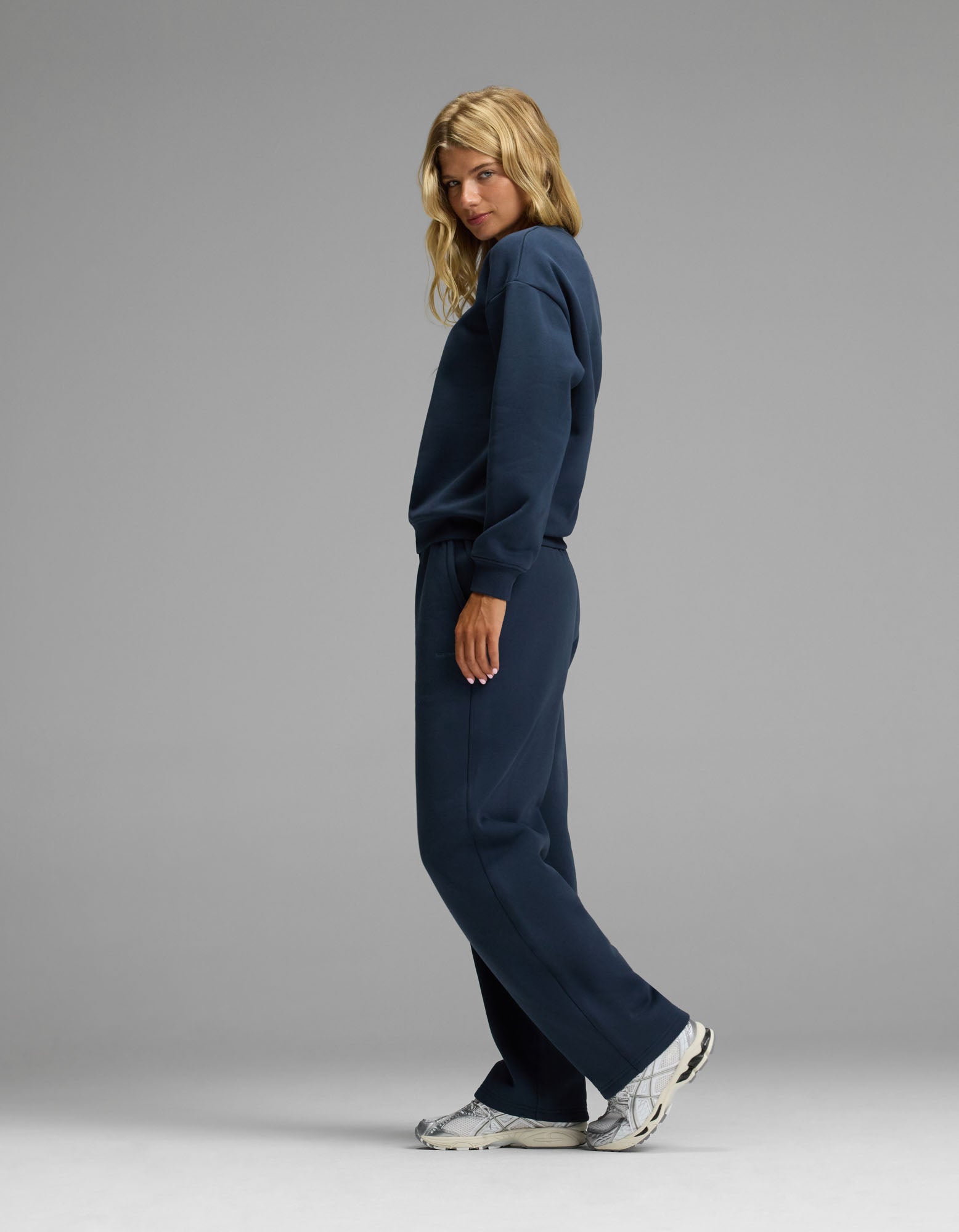 A person stands sideways, looking at the camera, wearing the Cozy Earth Women’s Cityscape Crewneck & Wide Leg Pant Set in navy blue with white sneakers, against a plain gray background.