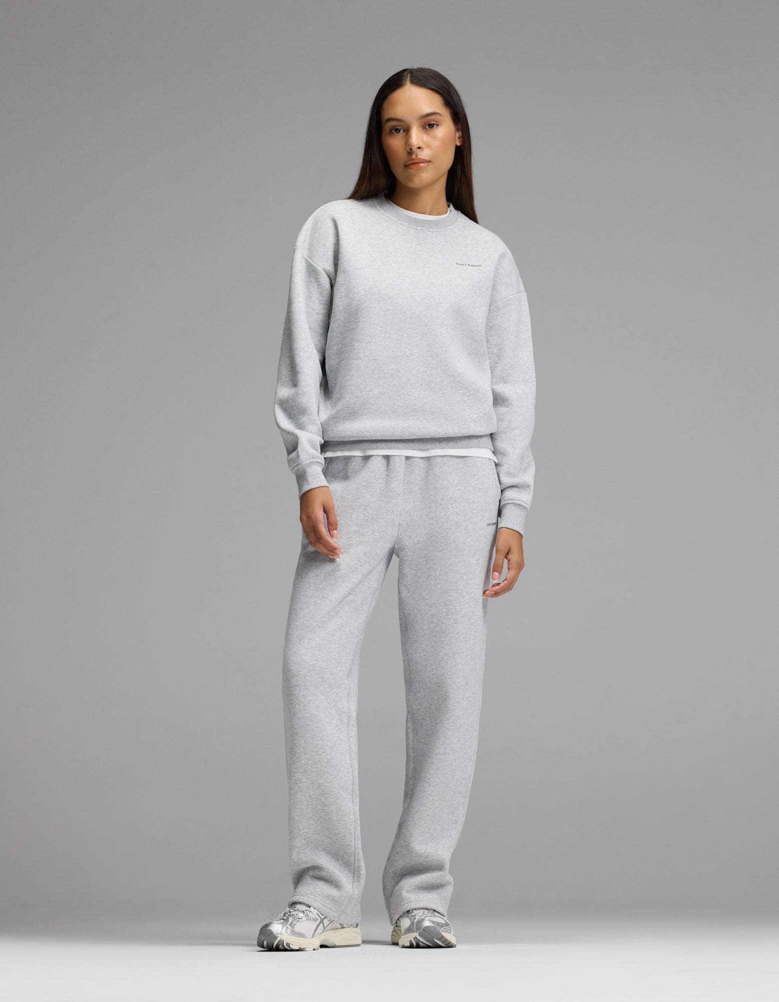 A woman stands facing forward against a plain gray background, wearing Cozy Earth’s Women’s Cityscape Crewneck & Wide Leg Pant Set in Heather Grey, paired with silver sneakers.|Color:Heather Grey