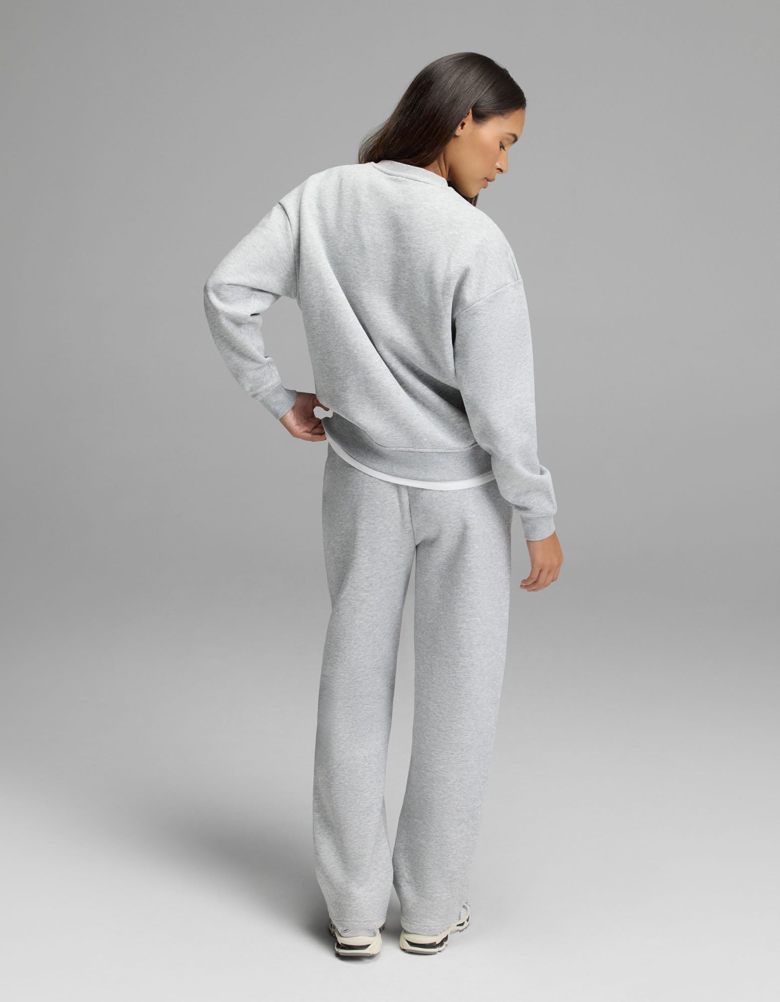 A person stands facing away, dressed in the Cozy Earth Women’s Cityscape Crewneck & Wide Leg Pant Set in light gray with white sneakers, against a plain gray background.|Color:Heather Grey