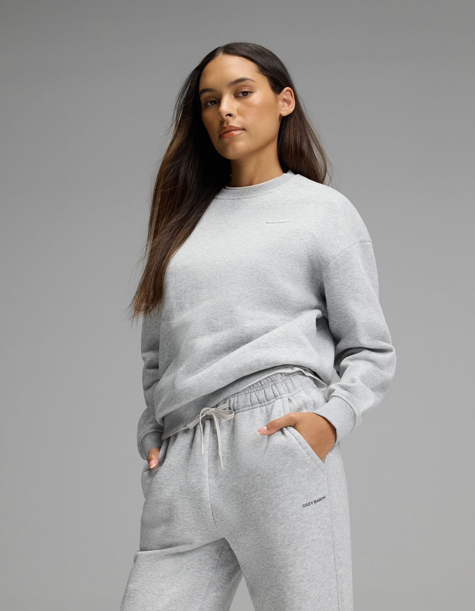 A woman with long brown hair models the Cozy Earth Women’s Cityscape Crewneck & Wide Leg Pant Set in light gray, standing with one hand in her pocket against a plain gray background.