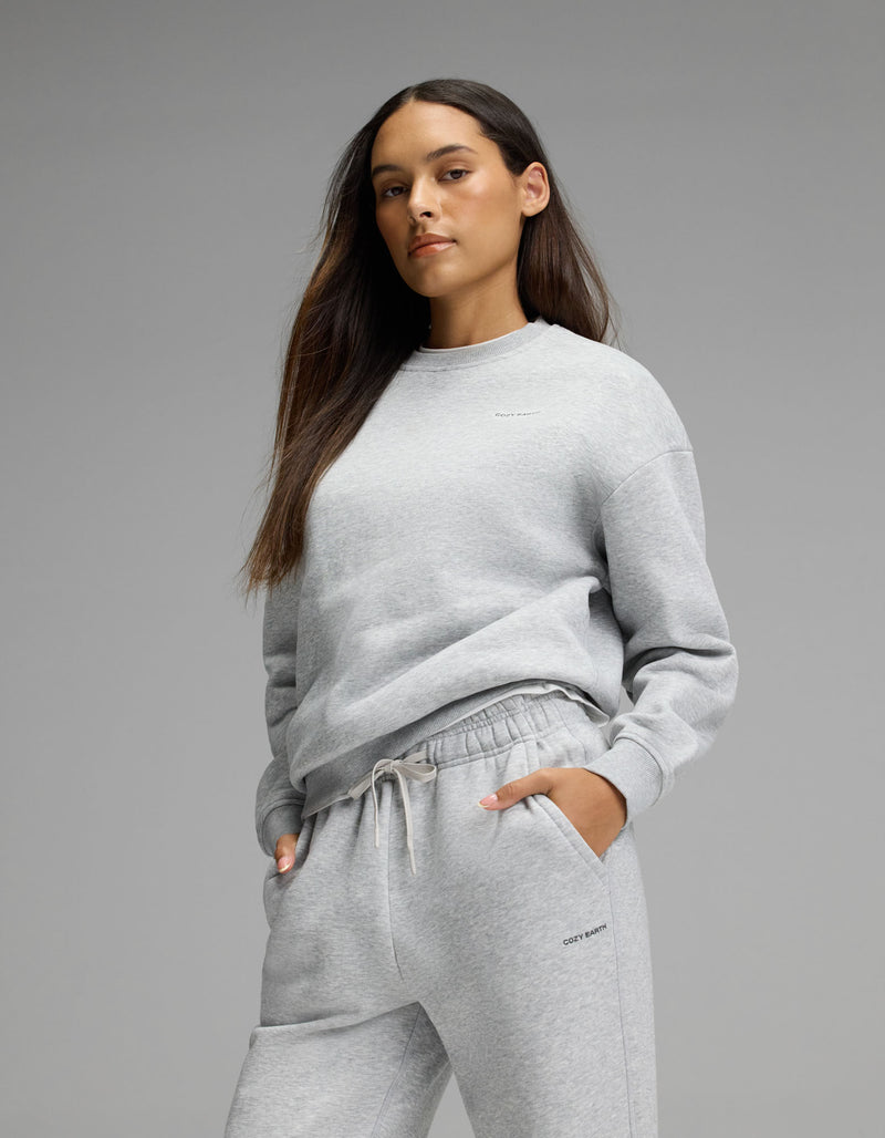 A woman with long brown hair models the Cozy Earth Women’s Cityscape Crewneck & Wide Leg Pant Set in light gray, standing with one hand in her pocket against a plain gray background.