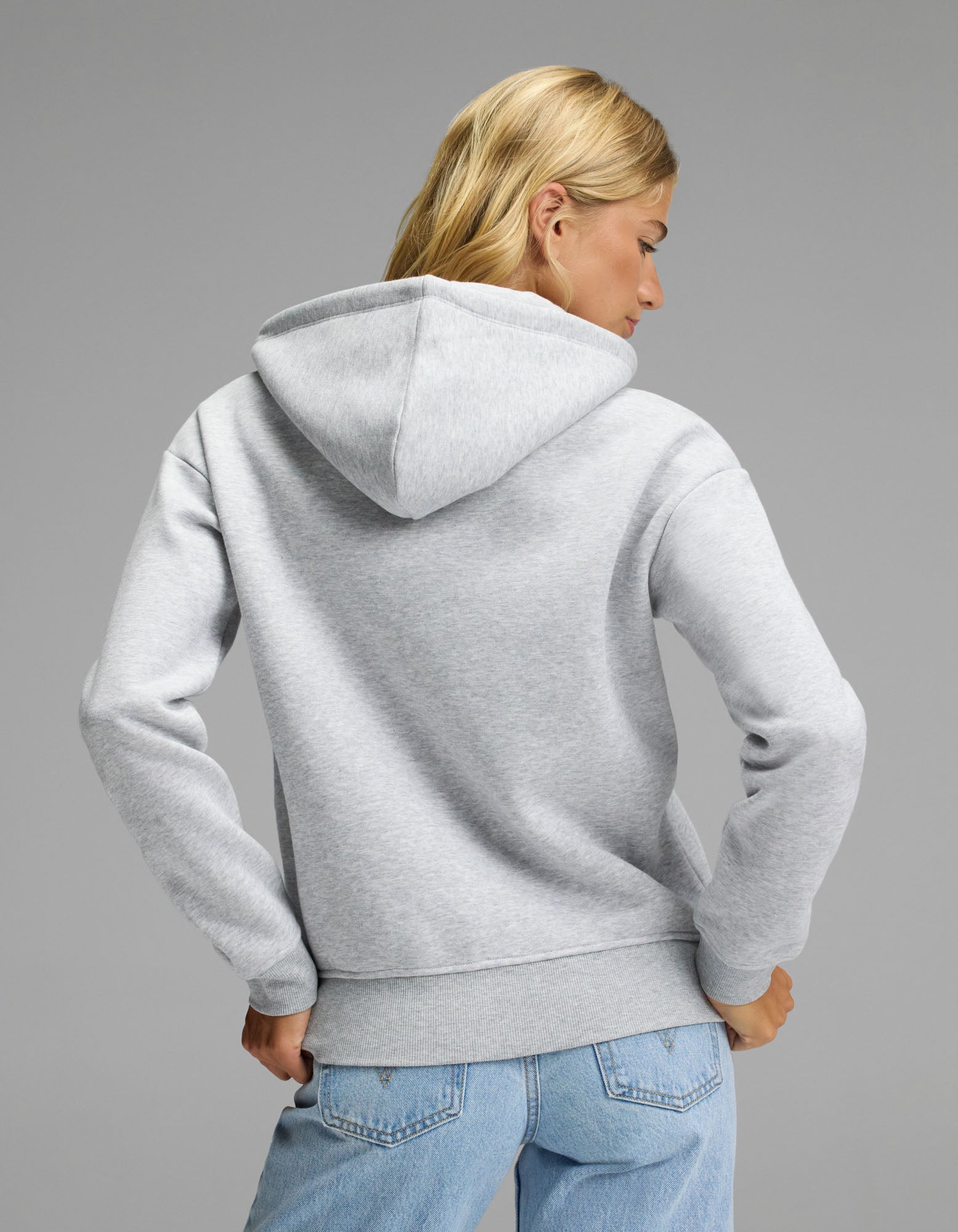 A woman with blonde hair, wearing the Cozy Earth Women’s CityScape Full Zip Hoodie in light gray and blue jeans, stands facing away from the camera against a plain gray background.
