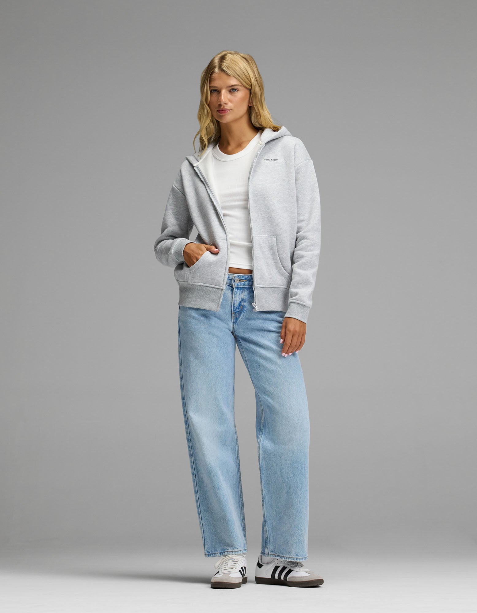 A woman with blonde hair and a neutral expression stands against a plain background, wearing the Cozy Earth Women’s CityScape Full Zip Hoodie in gray over a white t-shirt, paired with light blue jeans and white Adidas sneakers featuring black stripes.