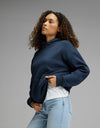 Wearing the Cozy Earth Women’s CityScape Hoodie in Eclipse over a white shirt and light blue jeans, a person with curly hair stands against a plain gray background, gazing slightly upward.|Color:Eclipse