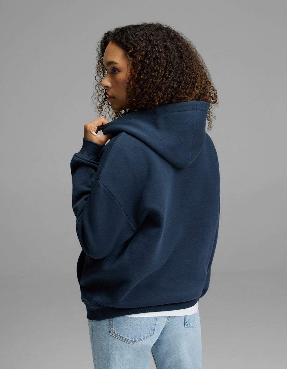 A woman with curly hair, dressed in the Cozy Earth Women’s CityScape Hoodie in navy blue and light blue jeans, stands facing away and gazes to the side against a plain gray background.
