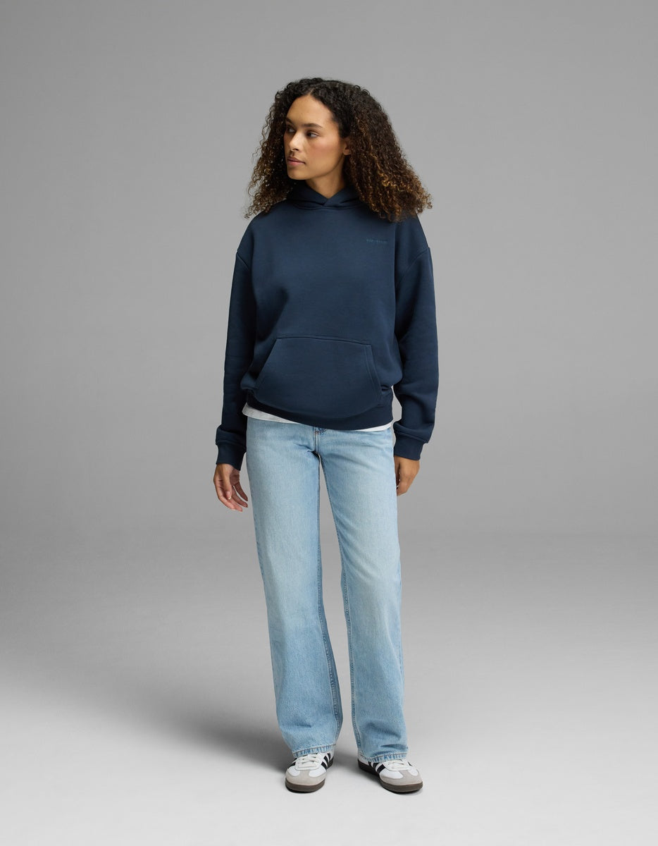 A person with curly hair is wearing the Cozy Earth Women’s CityScape Hoodie in navy blue, paired with light blue jeans and white sneakers, standing against a plain gray background and looking to the side with a neutral expression.