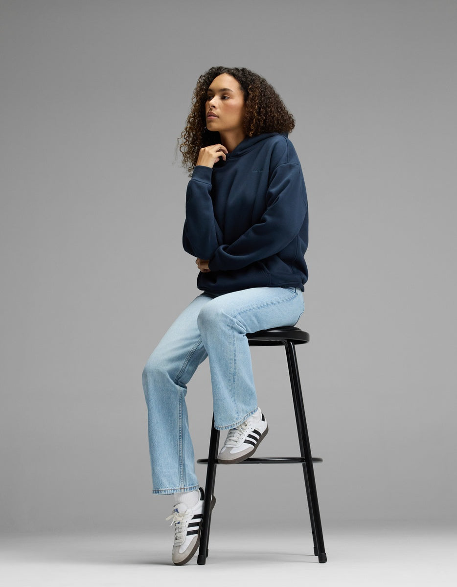 A person with curly hair sits on a tall black stool, wearing a Cozy Earth Women’s CityScape Hoodie in navy, light blue jeans, and white Adidas sneakers, against a plain gray background.
