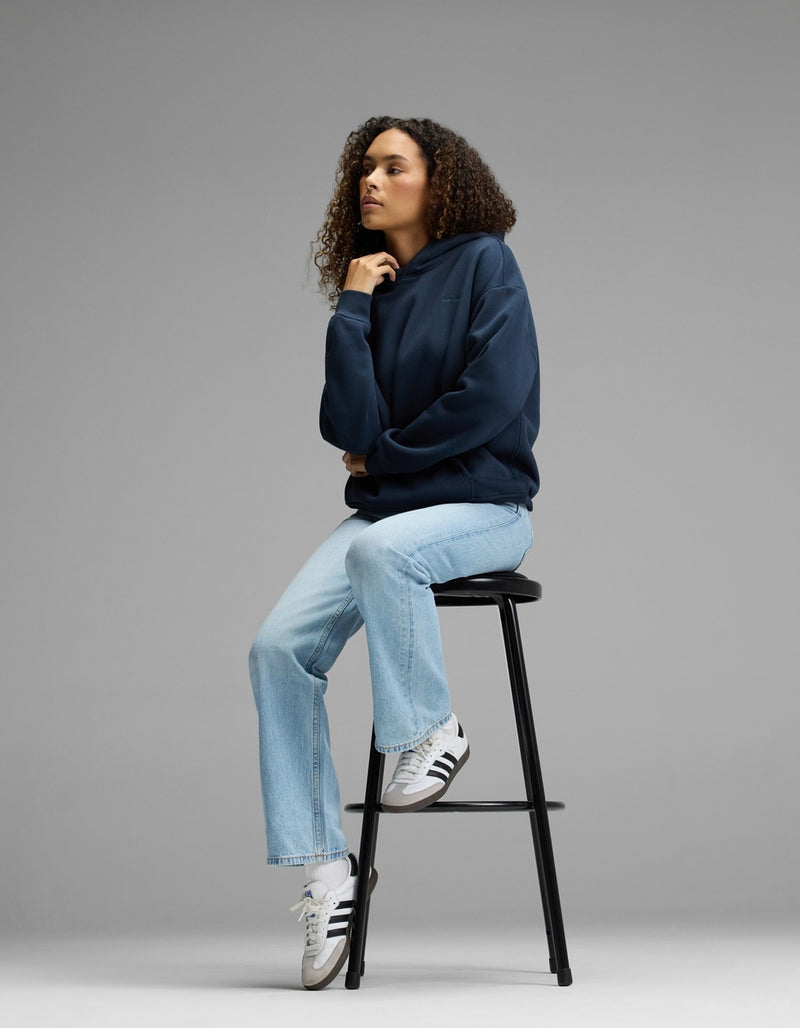 A person with curly hair sits on a tall black stool, wearing a Cozy Earth Women’s CityScape Hoodie in navy, light blue jeans, and white Adidas sneakers, against a plain gray background.
