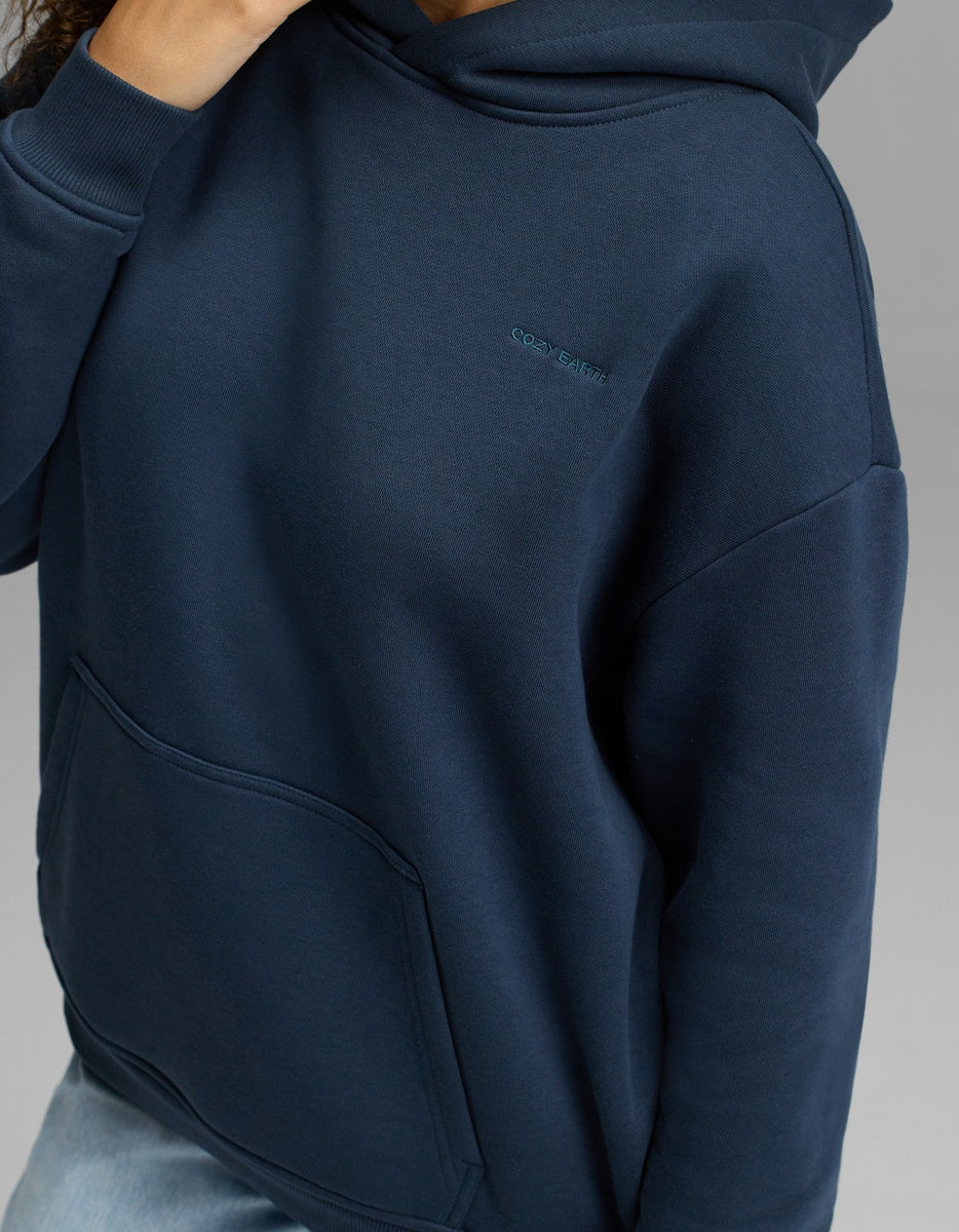 A person is wearing the Cozy Earth Women’s CityScape Hoodie in navy blue, featuring a front pocket and subtle embroidered text on the chest. Their hand rests near their neck against a plain gray background.