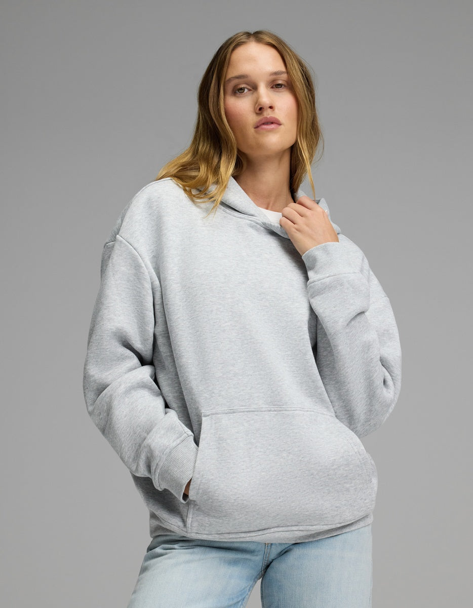 A woman models the Cozy Earth Women’s CityScape Hoodie in Heather Grey, pairing it with light blue jeans as she stands against a plain gray background—one hand adjusting the hoodie’s collar, the other tucked into its front pocket.|Color:Heather Grey
