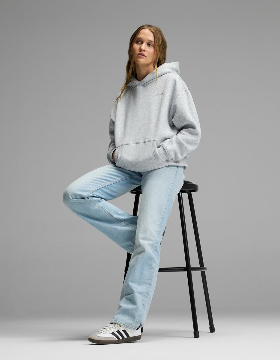 A woman wearing the Cozy Earth Women’s CityScape Hoodie and light blue jeans sits on a black stool against a plain gray background, gazing slightly to the side with a relaxed expression.