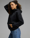 A person with curly hair poses in the Cozy Earth Women’s CityScape Hoodie in Jet Black and blue jeans against a plain gray background, one hand in the hoodie pocket and the other touching their hair.|Color:Jet Black