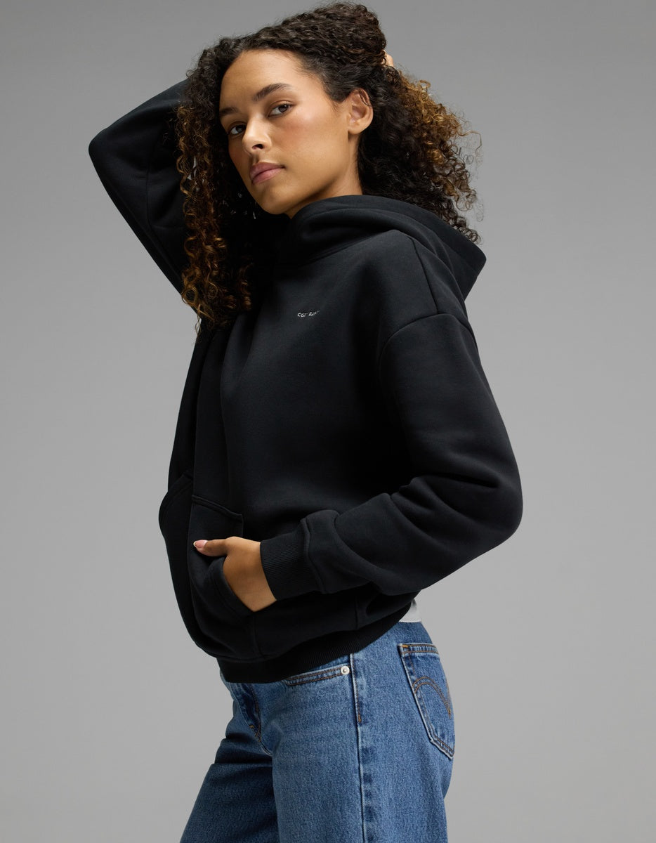 A person with curly hair poses in the Cozy Earth Women’s CityScape Hoodie in Jet Black and blue jeans against a plain gray background, one hand in the hoodie pocket and the other touching their hair.|Color:Jet Black