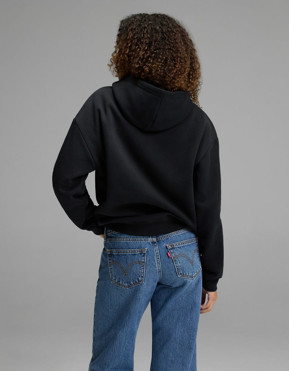 A woman with curly hair is seen from behind, wearing the Cozy Earth Women’s CityScape Hoodie in black and blue jeans, standing against a plain gray backdrop.