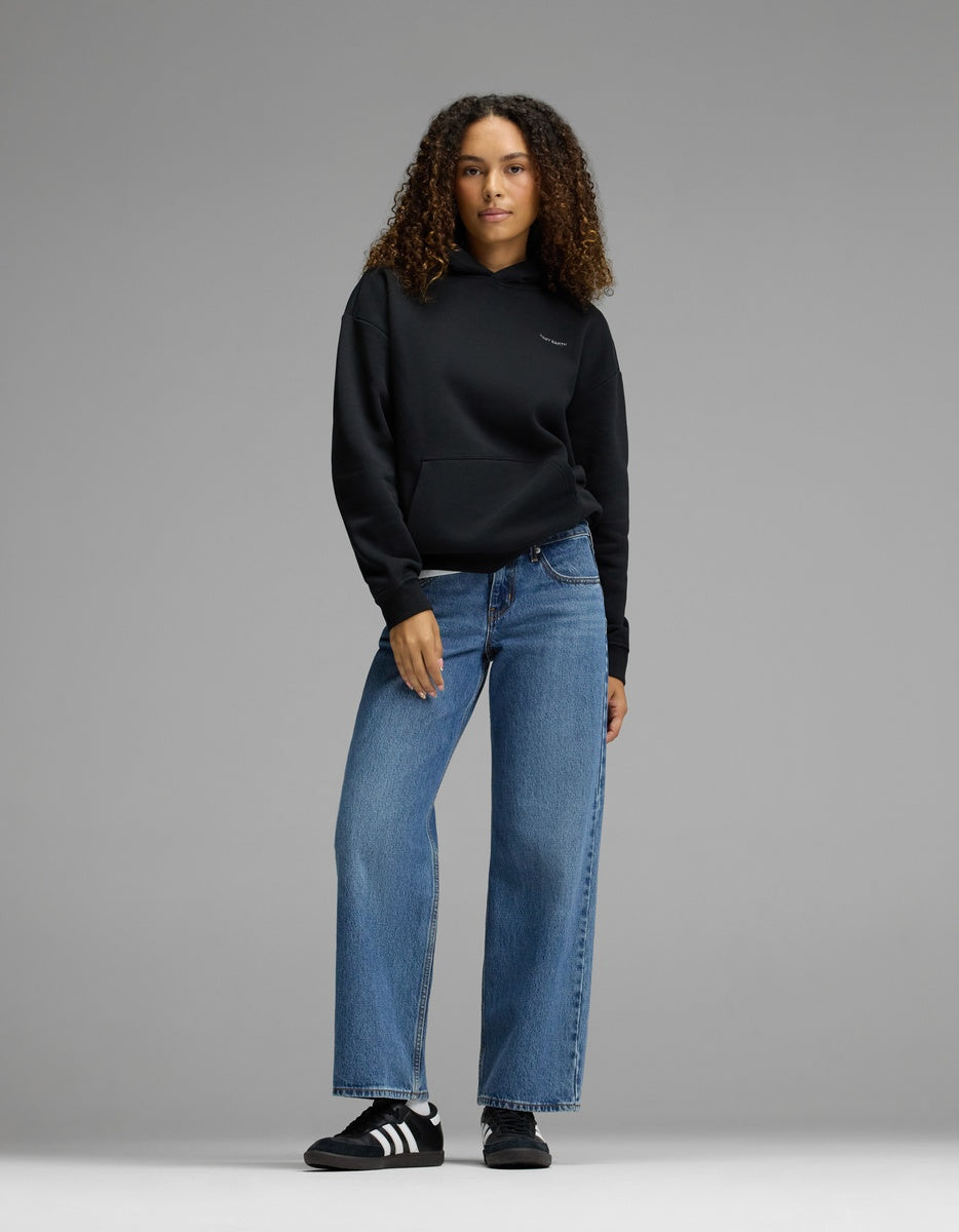 A woman with curly hair wears the Cozy Earth Women’s CityScape Hoodie in black, paired with blue wide-leg jeans and black-and-white sneakers, standing against a plain gray background.