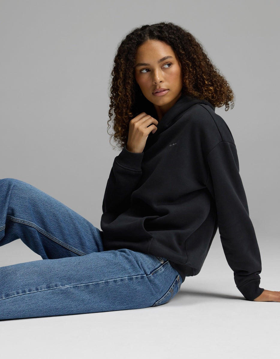 A woman with curly hair sits on the floor, wearing the Cozy Earth Women’s CityScape Hoodie and blue jeans, looking to the side against a plain gray background.