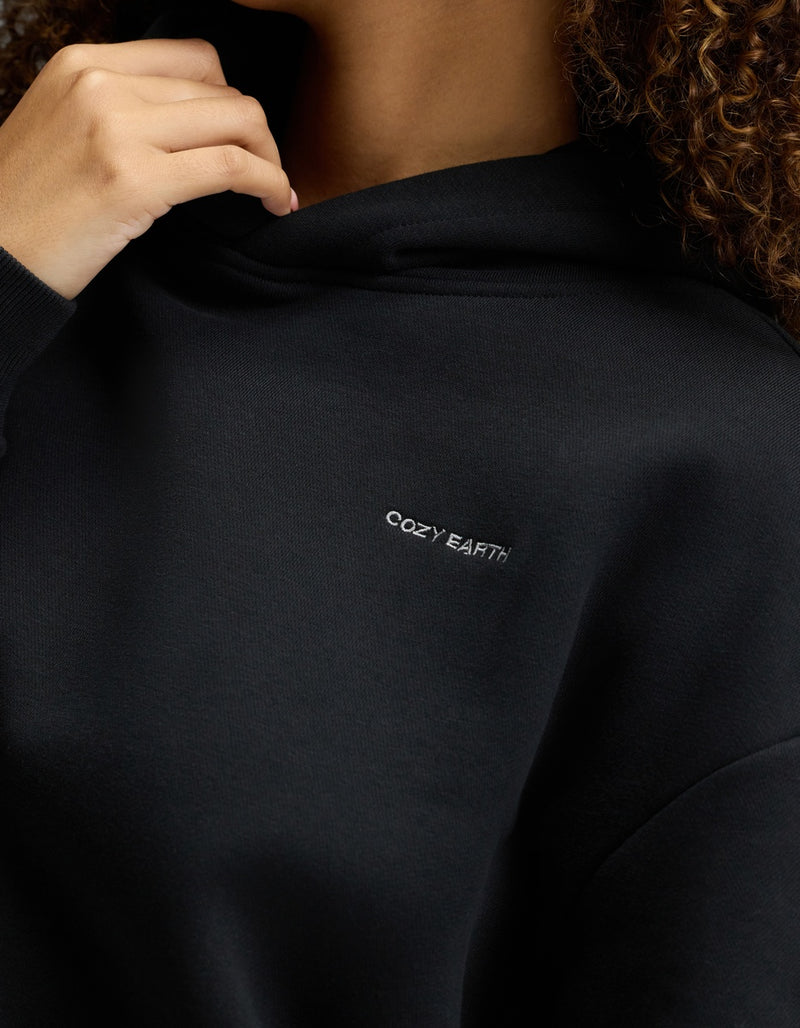 A person wearing the Cozy Earth Women’s CityScape Hoodie in black, featuring “COZY EARTH” embroidered in white on the chest, holds the hood near their face with only part of their face and hand visible.