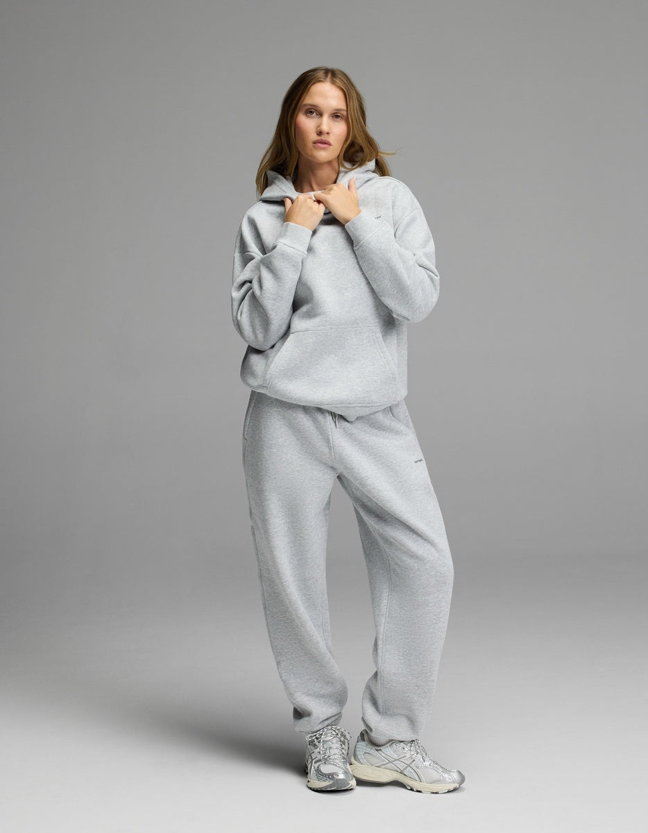 A woman stands against a grey background wearing the Cozy Earth Women’s CityScape Hoodie & Sweatpant Set, paired with silver sneakers. She holds the strings of her hoodie and looks at the camera.|Color:Heather Grey