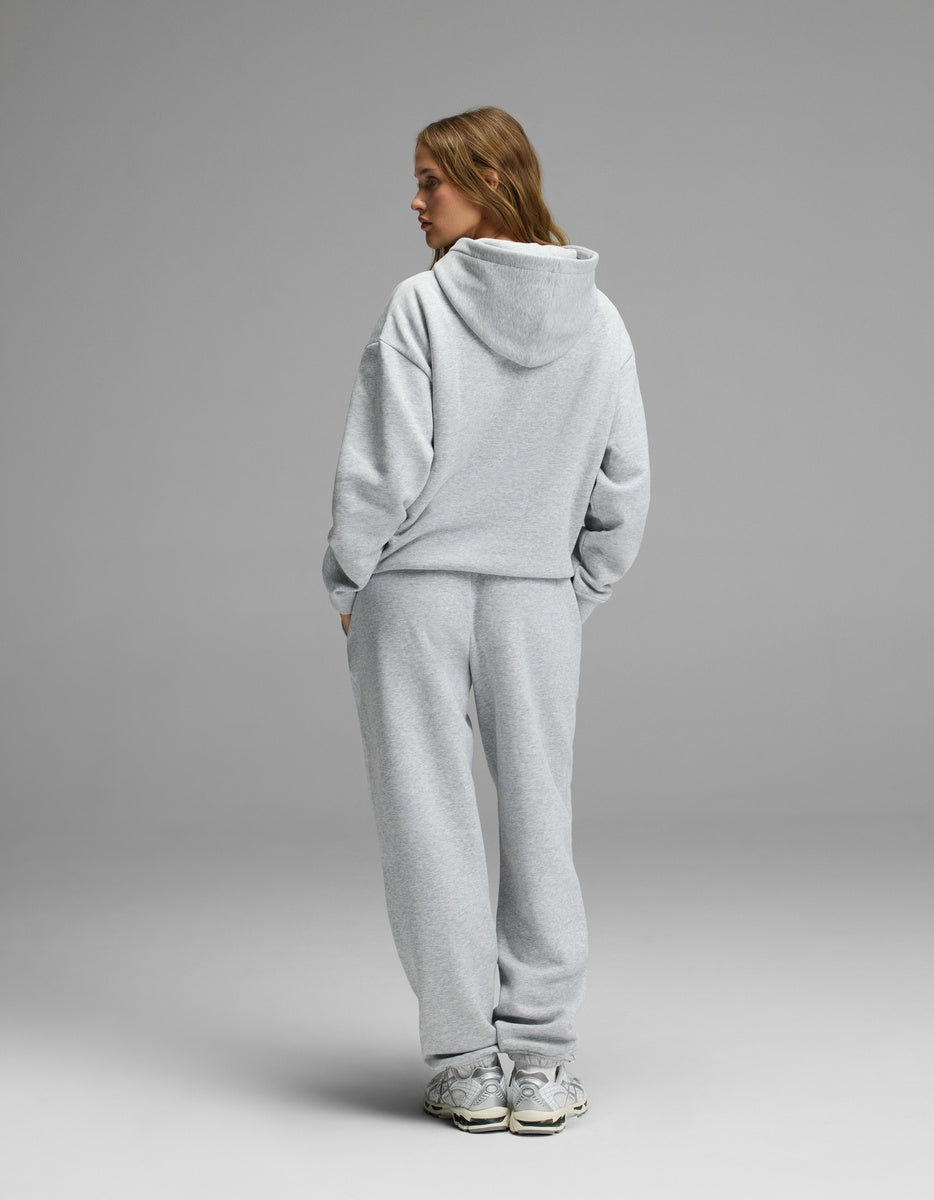 A woman models the Cozy Earth Women’s CityScape Hoodie & Sweatpant Set in light gray, standing with her back to the camera and hands in her pockets against a plain gray background.