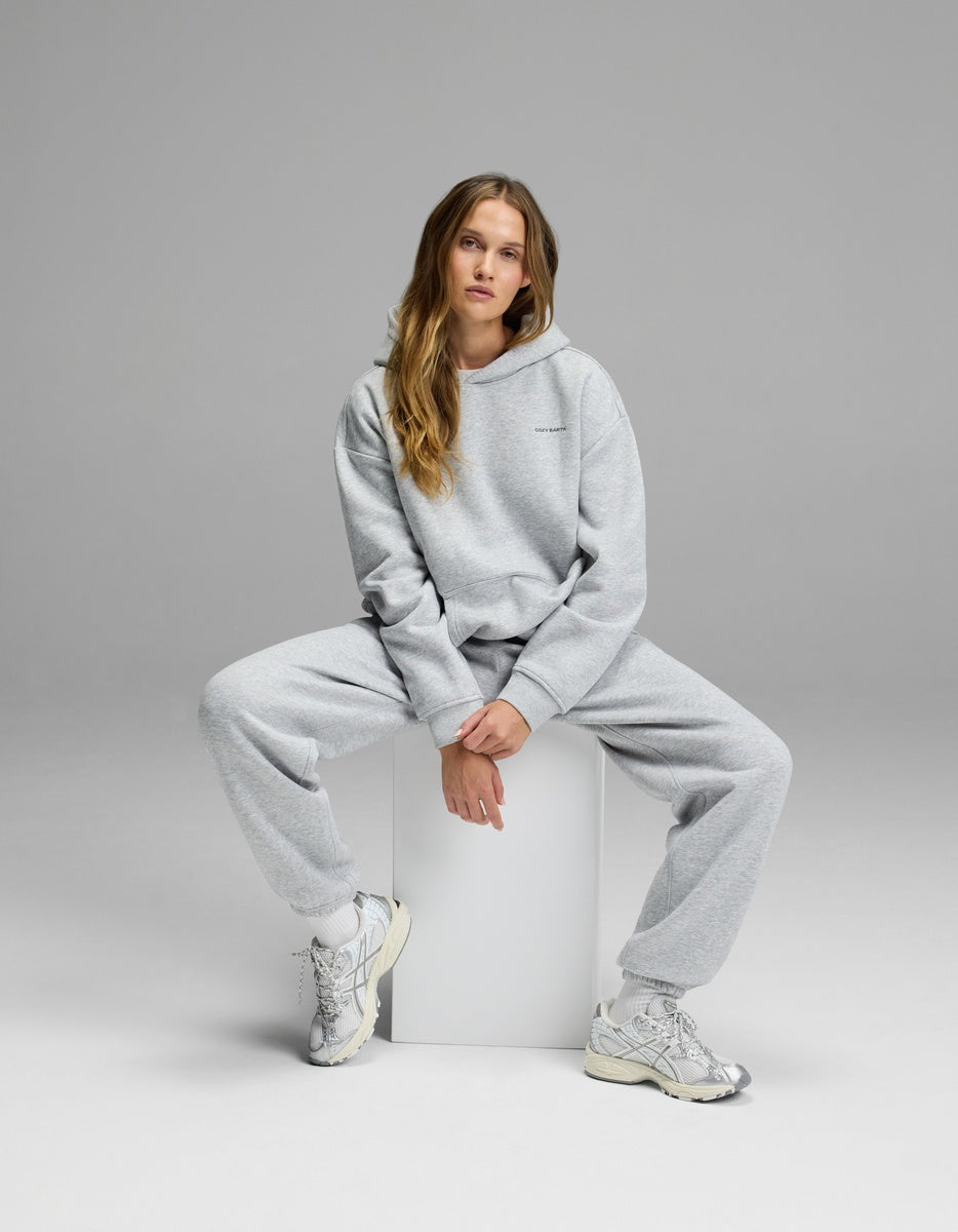 Wearing the Cozy Earth Women’s CityScape Hoodie & Sweatpant Set, a woman sits on a white cube against a plain gray background in white sneakers, gazing at the camera with a relaxed expression.