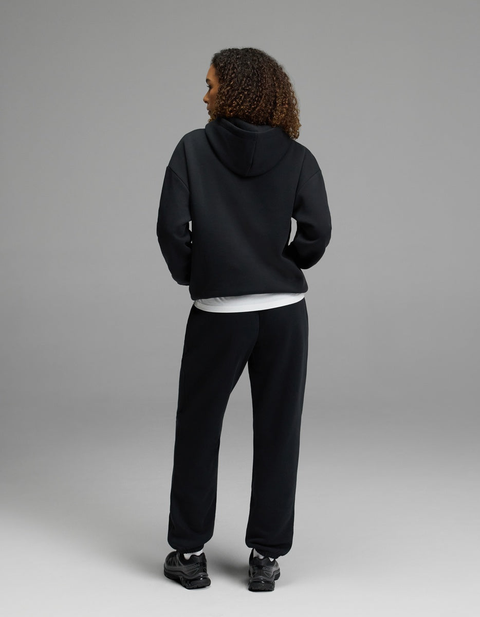 A woman stands with her back to the camera, wearing the black Women’s CityScape Hoodie & Sweatpant Set by Cozy Earth over a white shirt and paired with black sneakers, set against a plain gray background.