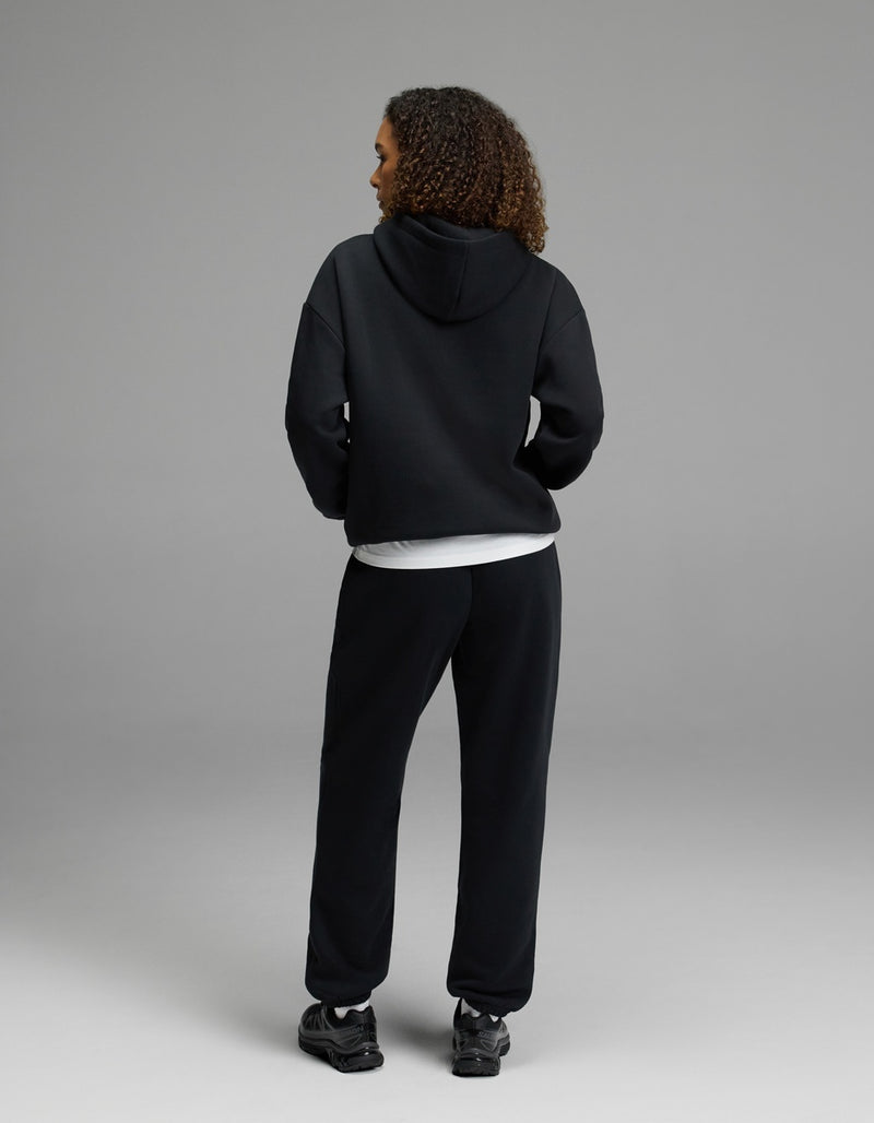 A woman stands with her back to the camera, wearing the black Women’s CityScape Hoodie & Sweatpant Set by Cozy Earth over a white shirt and paired with black sneakers, set against a plain gray background.