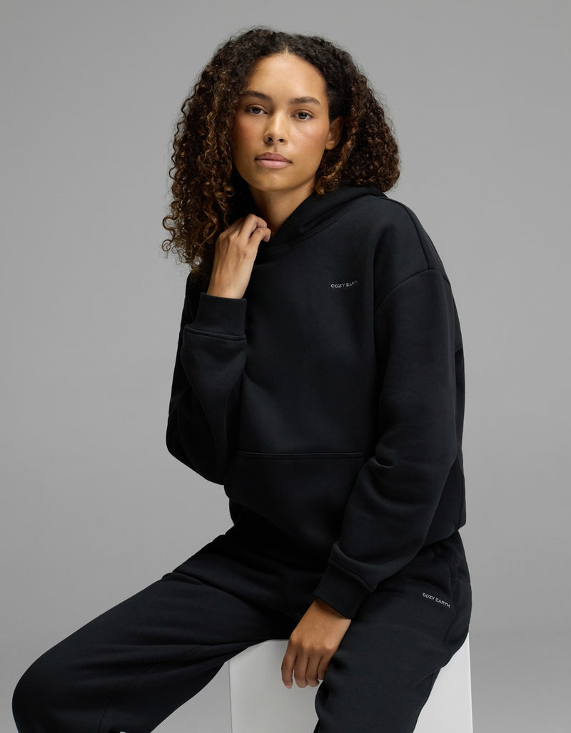 A woman with curly hair models the Cozy Earth Women’s CityScape Hoodie & Sweatpant Set in black, sitting on a white cube against a plain gray background and looking calmly at the camera.