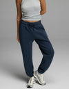 A person is shown from the shoulders down, standing against a plain gray background and wearing navy blue Cozy Earth Women’s CityScape Sweatpants, a grey sleeveless crop top, and white athletic shoes. Their head is not visible in the image.|Color:Eclipse