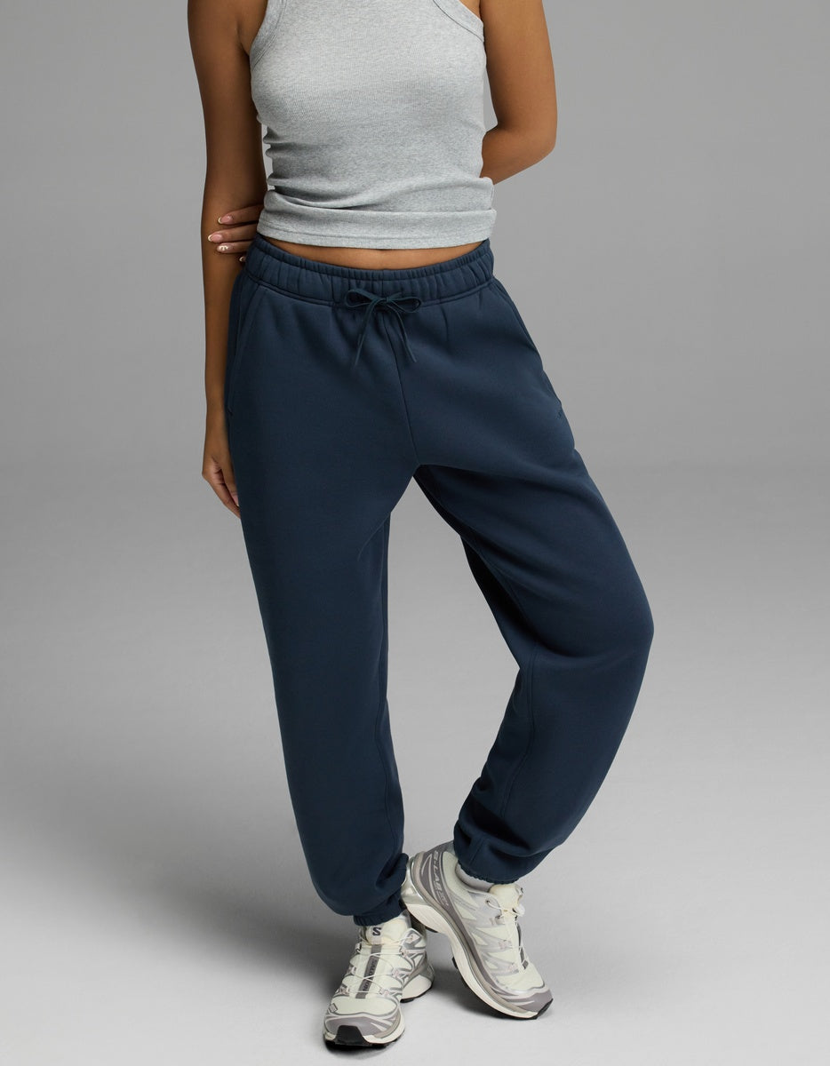 A person is shown from the shoulders down, standing against a plain gray background and wearing navy blue Cozy Earth Women’s CityScape Sweatpants, a grey sleeveless crop top, and white athletic shoes. Their head is not visible in the image.