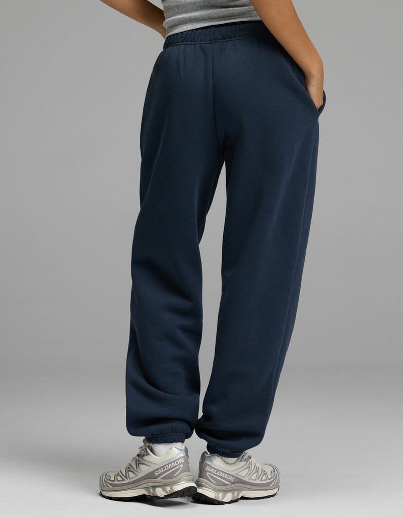 A person stands with their back to the camera, wearing a gray tank top, Cozy Earth Women’s CityScape Sweatpant in navy blue, and gray athletic shoes. One hand is in their pocket against a plain light gray background.