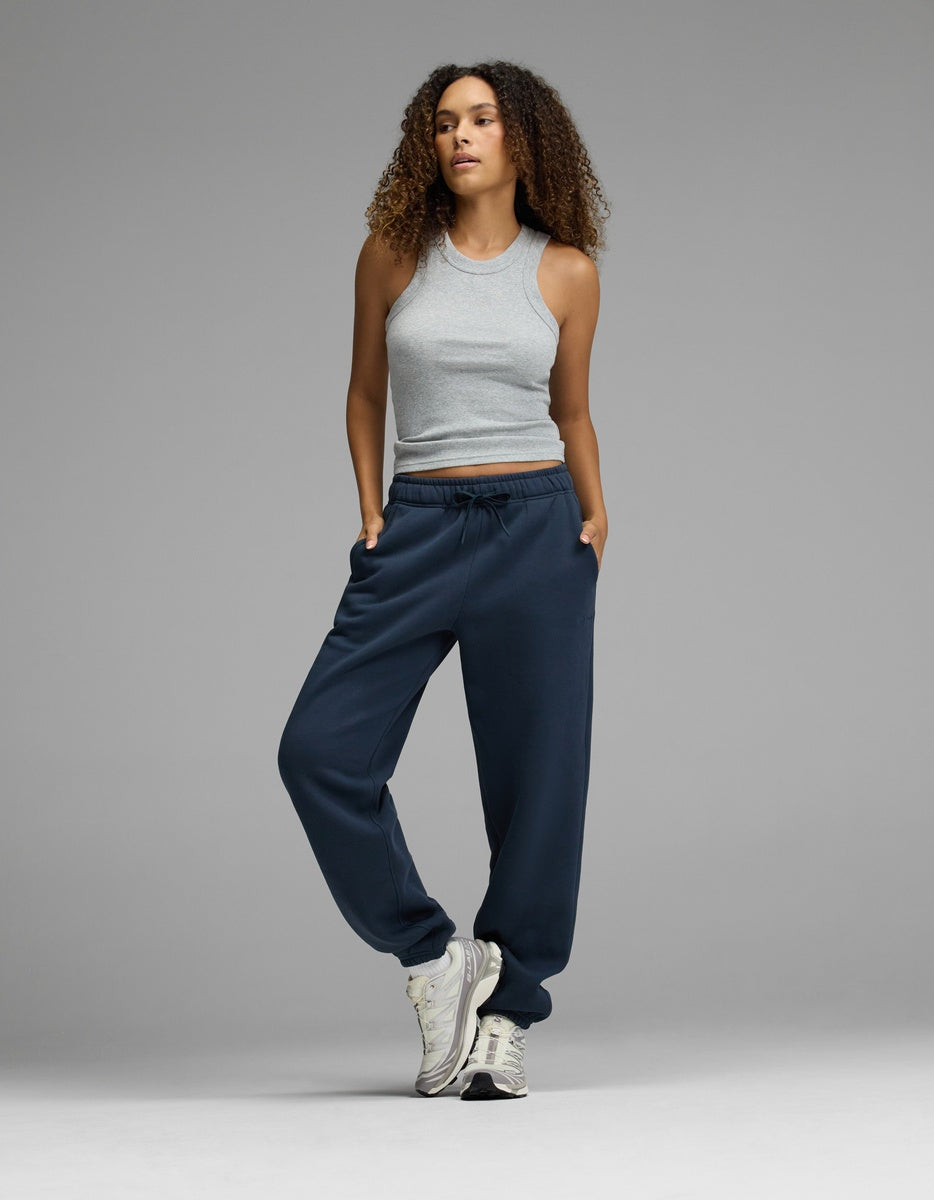 A woman with curly hair wears a gray sleeveless top, Cozy Earth’s Women’s CityScape Sweatpant in navy blue, and white sneakers. She stands with one foot slightly raised and her hands in her pockets against a plain gray background.