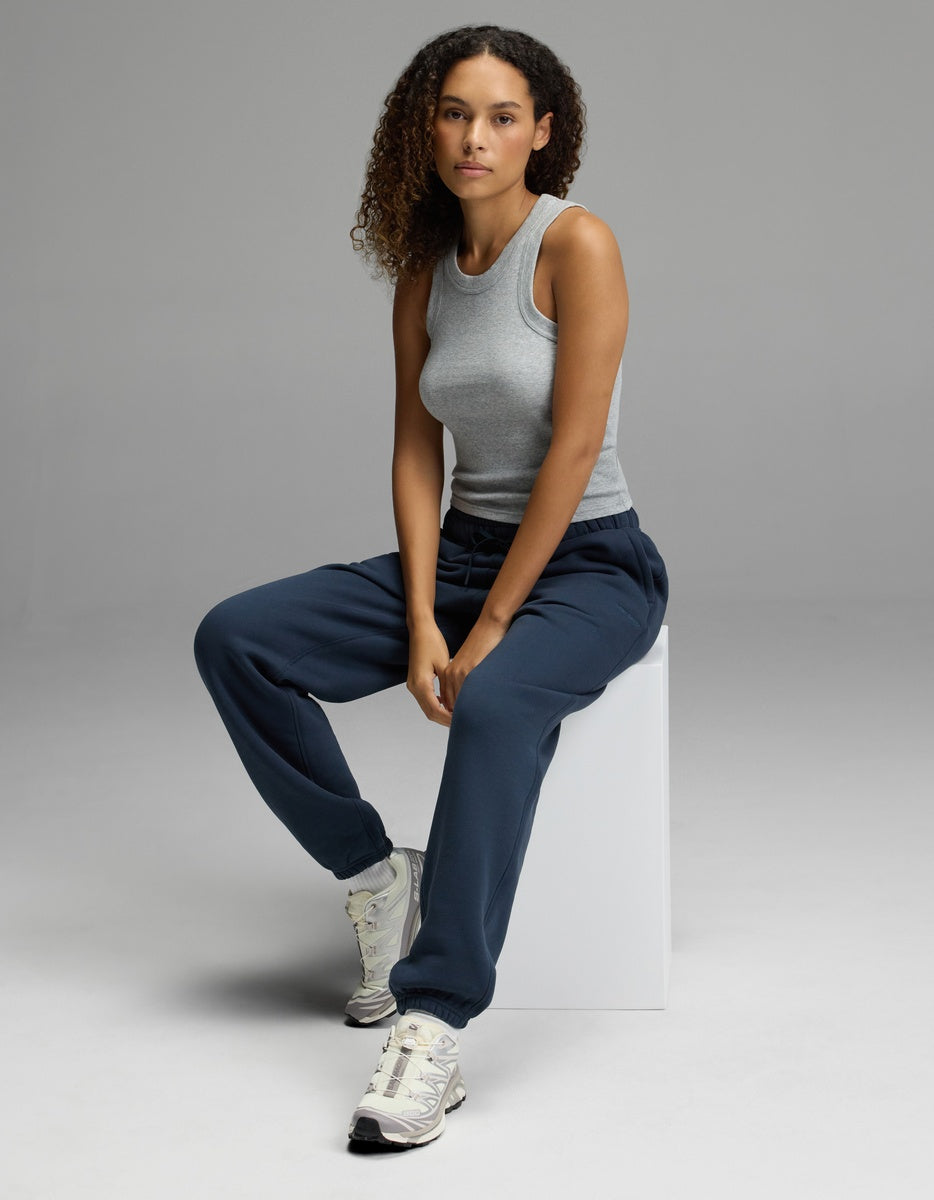 A person with curly hair, wearing a gray tank top, Cozy Earth Women’s CityScape Sweatpant in navy, and sneakers, sits on a white block against a plain gray background, looking at the camera.