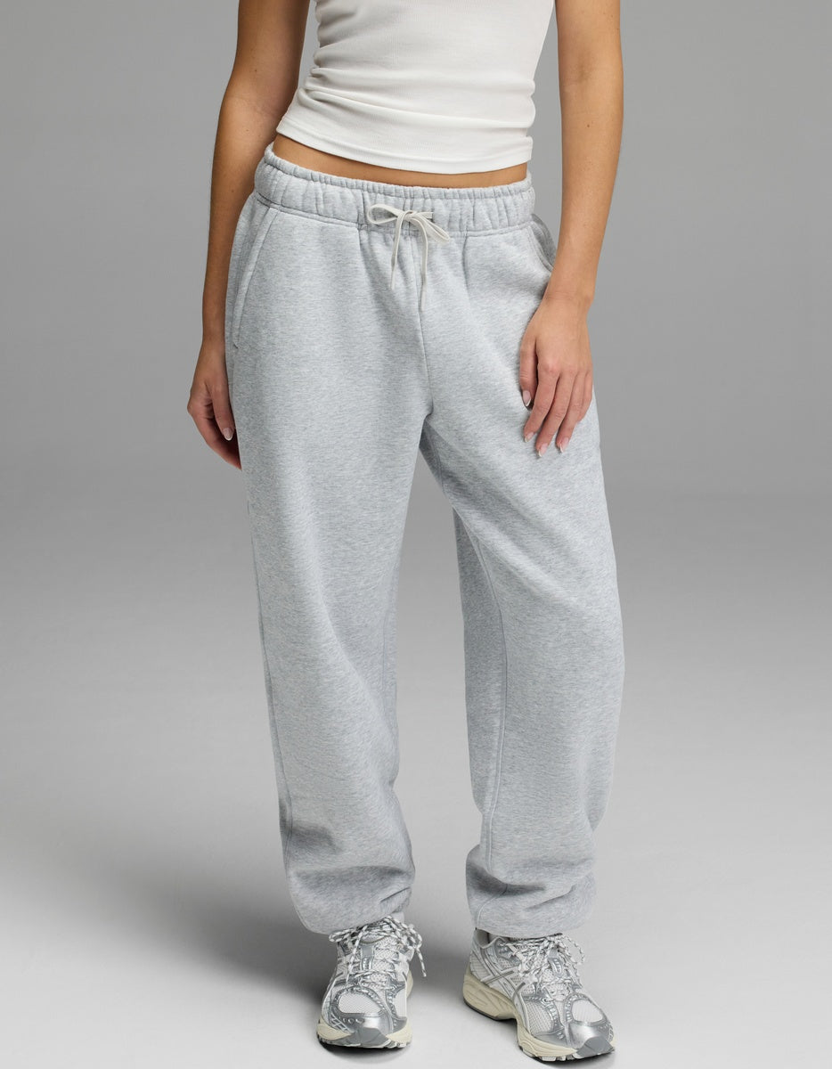 A person models the Cozy Earth Women’s CityScape Sweatpant in light gray, featuring a drawstring waist, paired with a white tank top and silver athletic shoes against a plain gray background. Only the lower half of the body is visible.|Color:Heather Grey