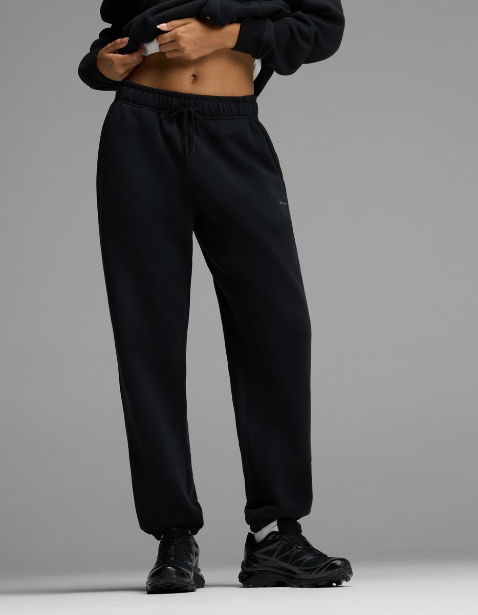 A person wearing Cozy Earth’s Women’s CityScape Sweatpant in black, paired with black sneakers and a cropped black hoodie, stands against a gray background with hands near the waistband.|Color:Jet Black