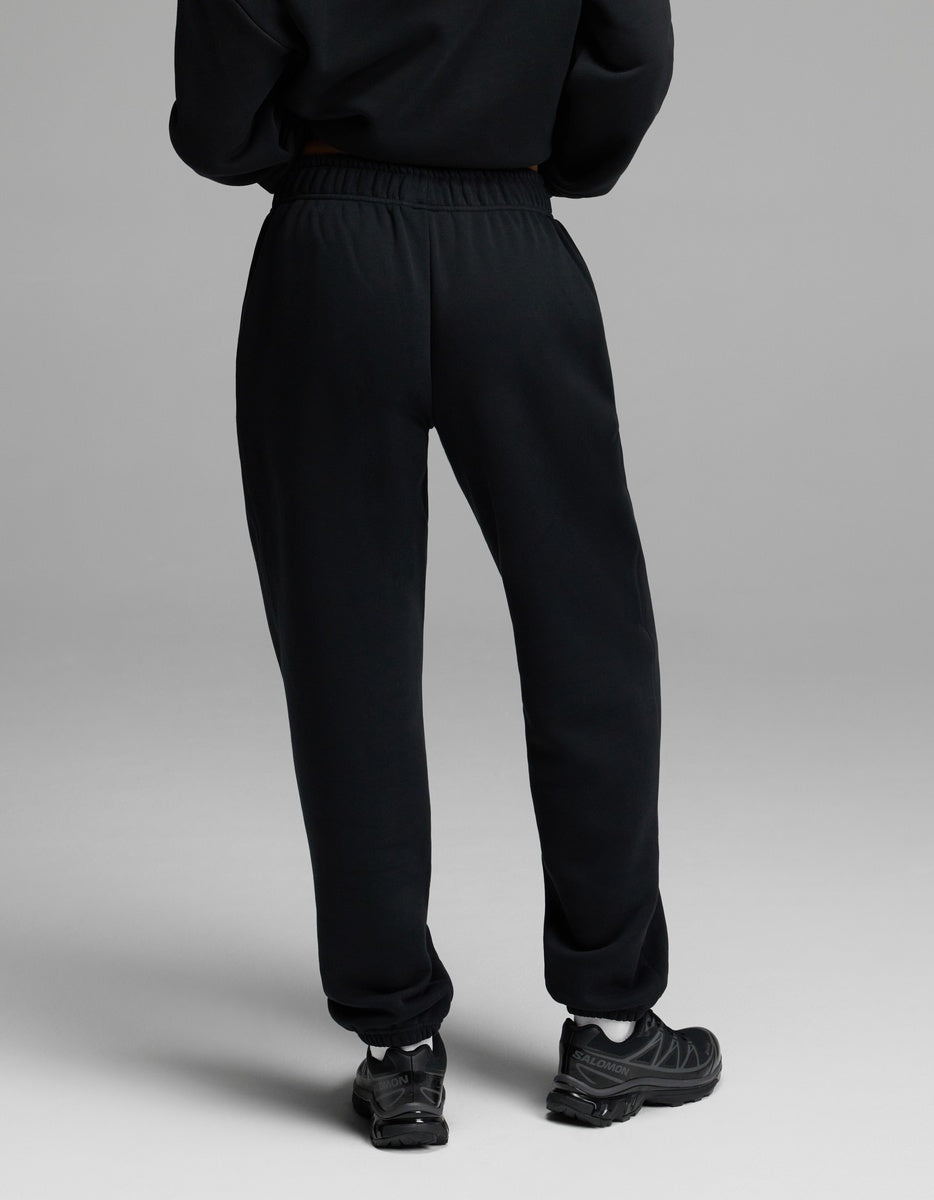 A model wears the Women’s CityScape Sweatpant by Cozy Earth in black with black sneakers, standing on a light grey background and photographed from behind.