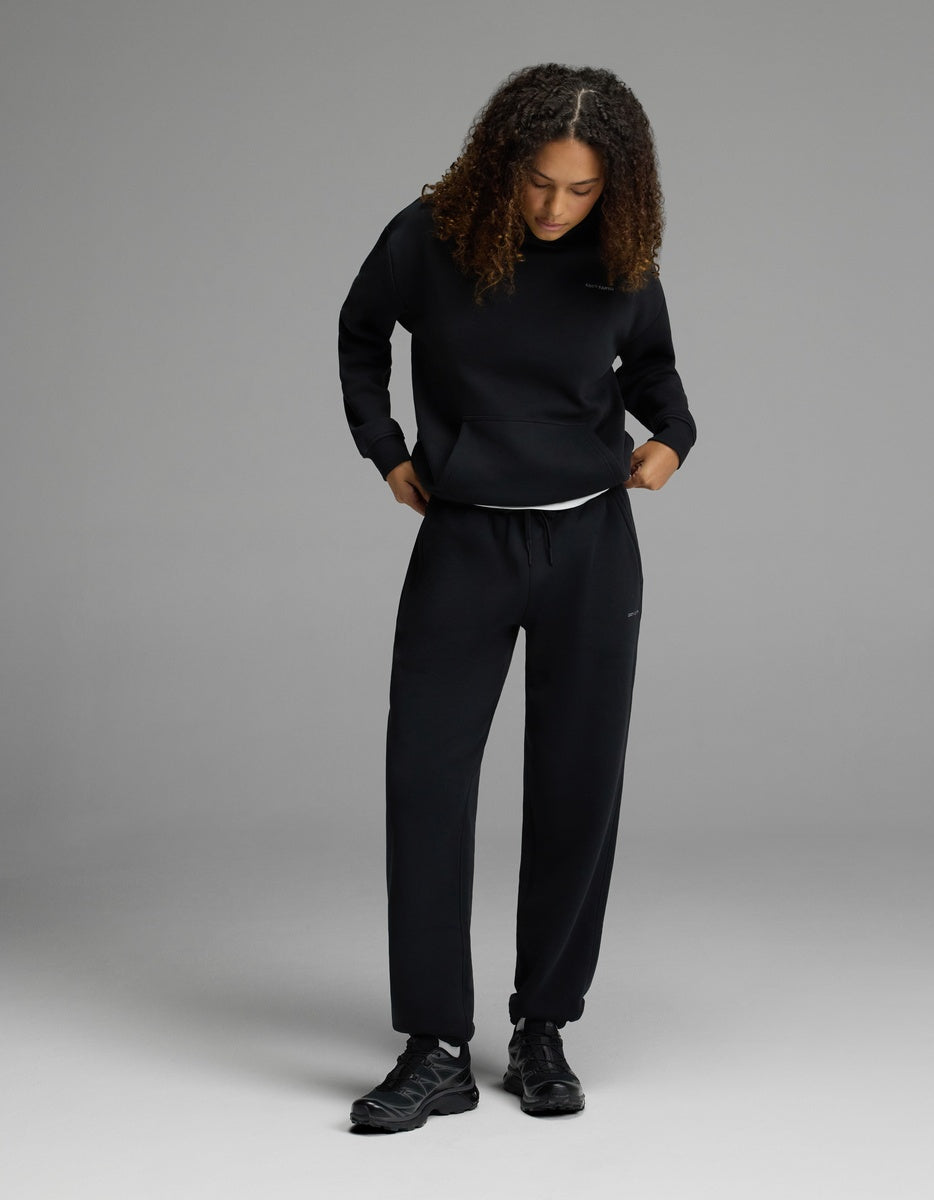 A person with curly hair wears Cozy Earth’s Women’s CityScape Sweatpant in black, paired with a black sweatshirt and black sneakers, standing on a plain light gray background and looking down with hands in pockets.