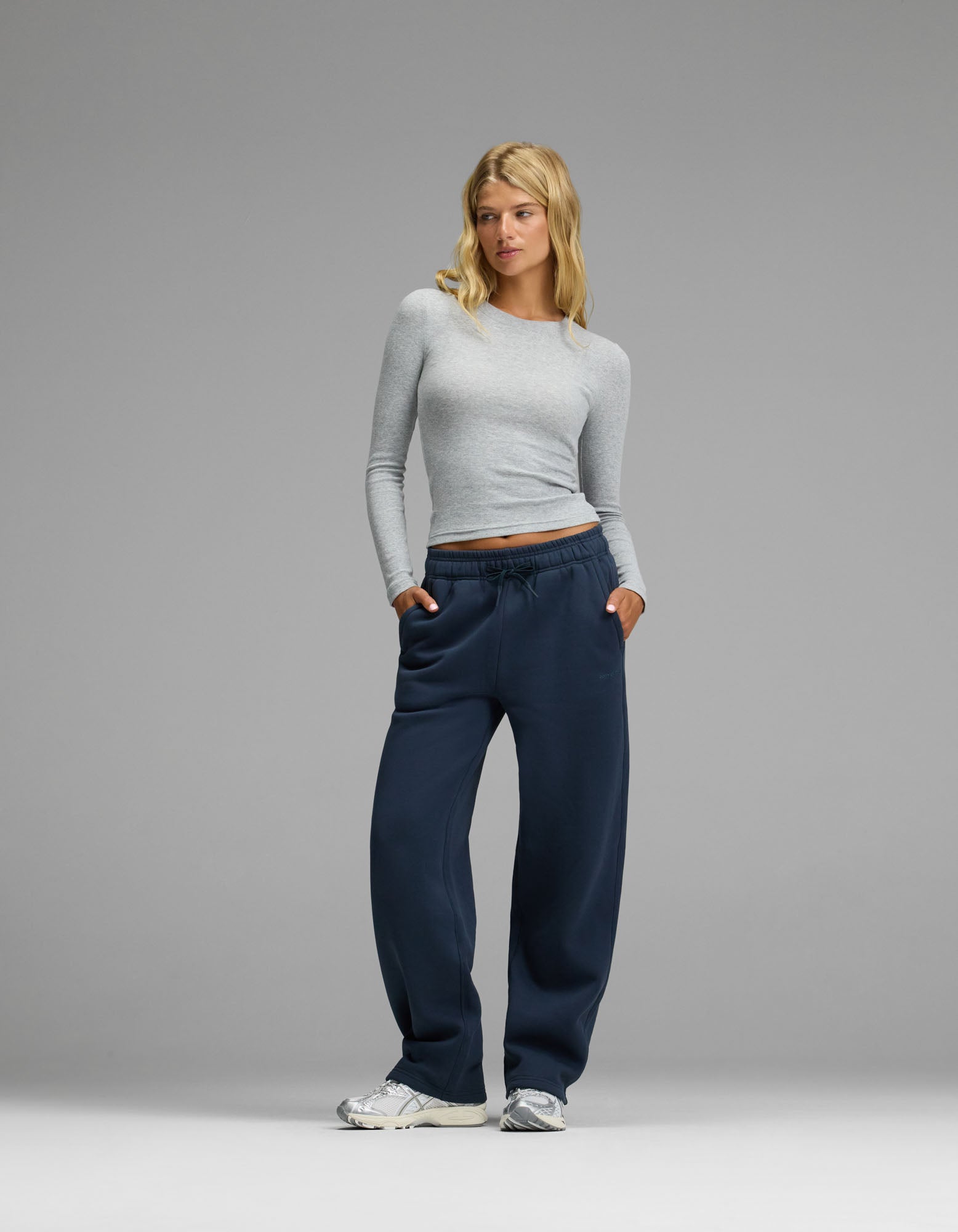A woman with blonde hair wearing a light gray long-sleeve shirt, Cozy Earth Women’s Cityscape Wide Leg Pants in navy blue, and white sneakers stands with her hands in her pockets against a plain gray background.