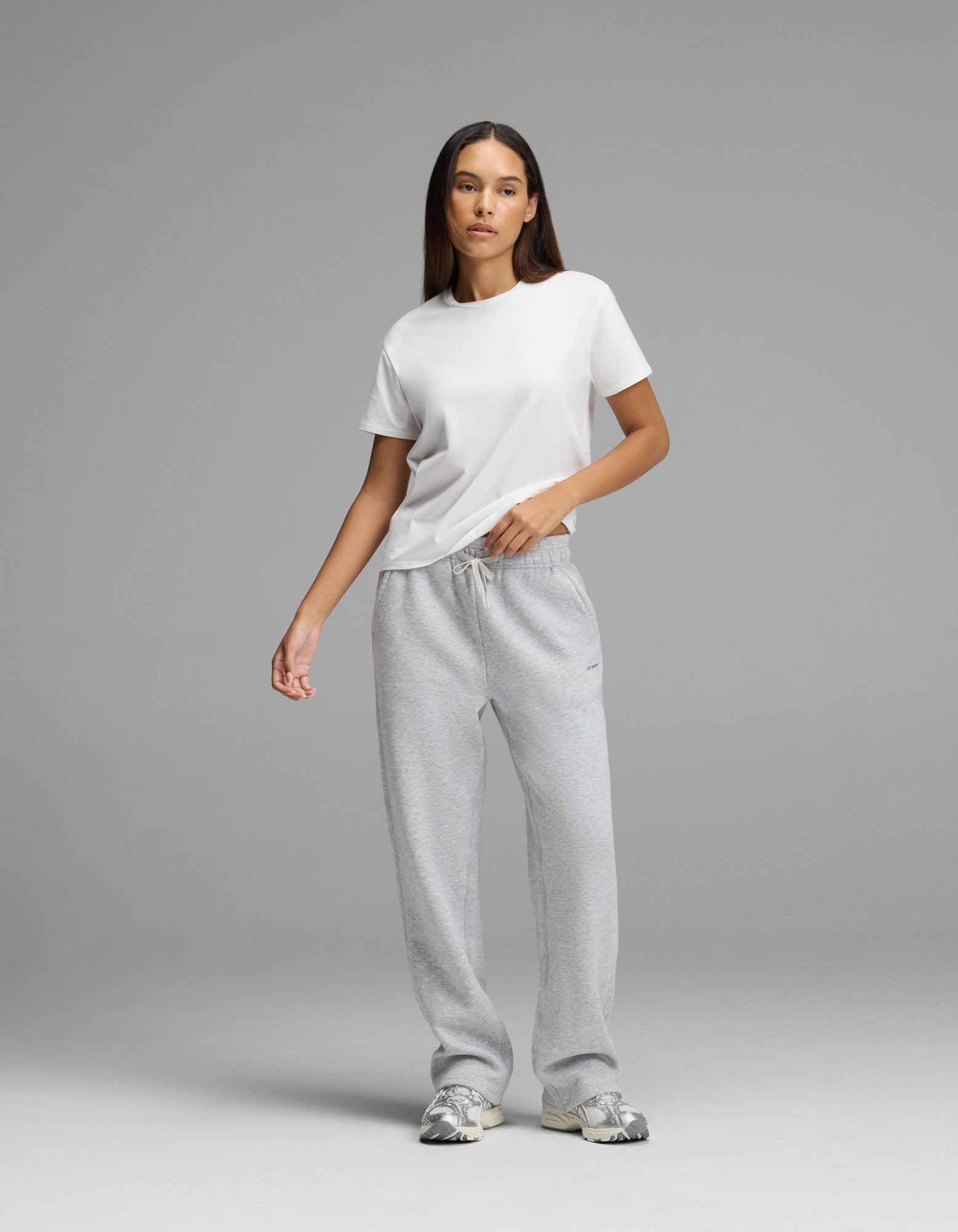 A person stands against a plain gray background wearing a white t-shirt, Cozy Earth Women’s Cityscape Wide Leg Pant in light gray, and gray sneakers. They have long, straight hair and a neutral expression with one hand in their pocket.