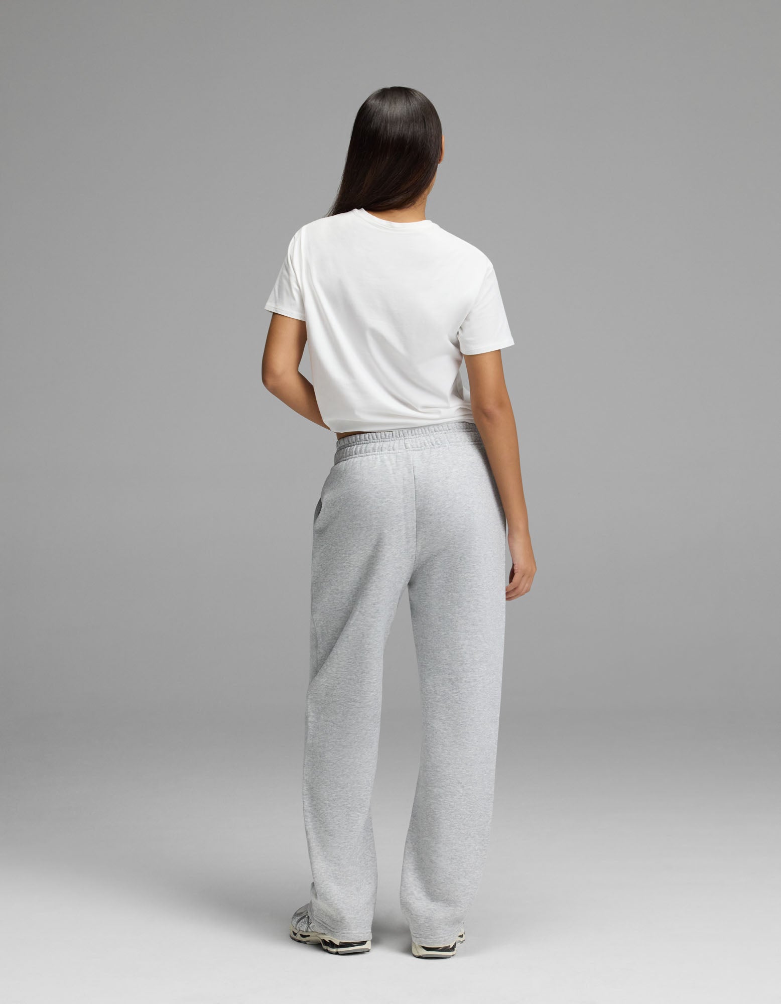 A person with long dark hair stands facing away, wearing a white t-shirt, Cozy Earth Women’s Cityscape Wide Leg Pant in light gray, and white sneakers against a plain gray background.
