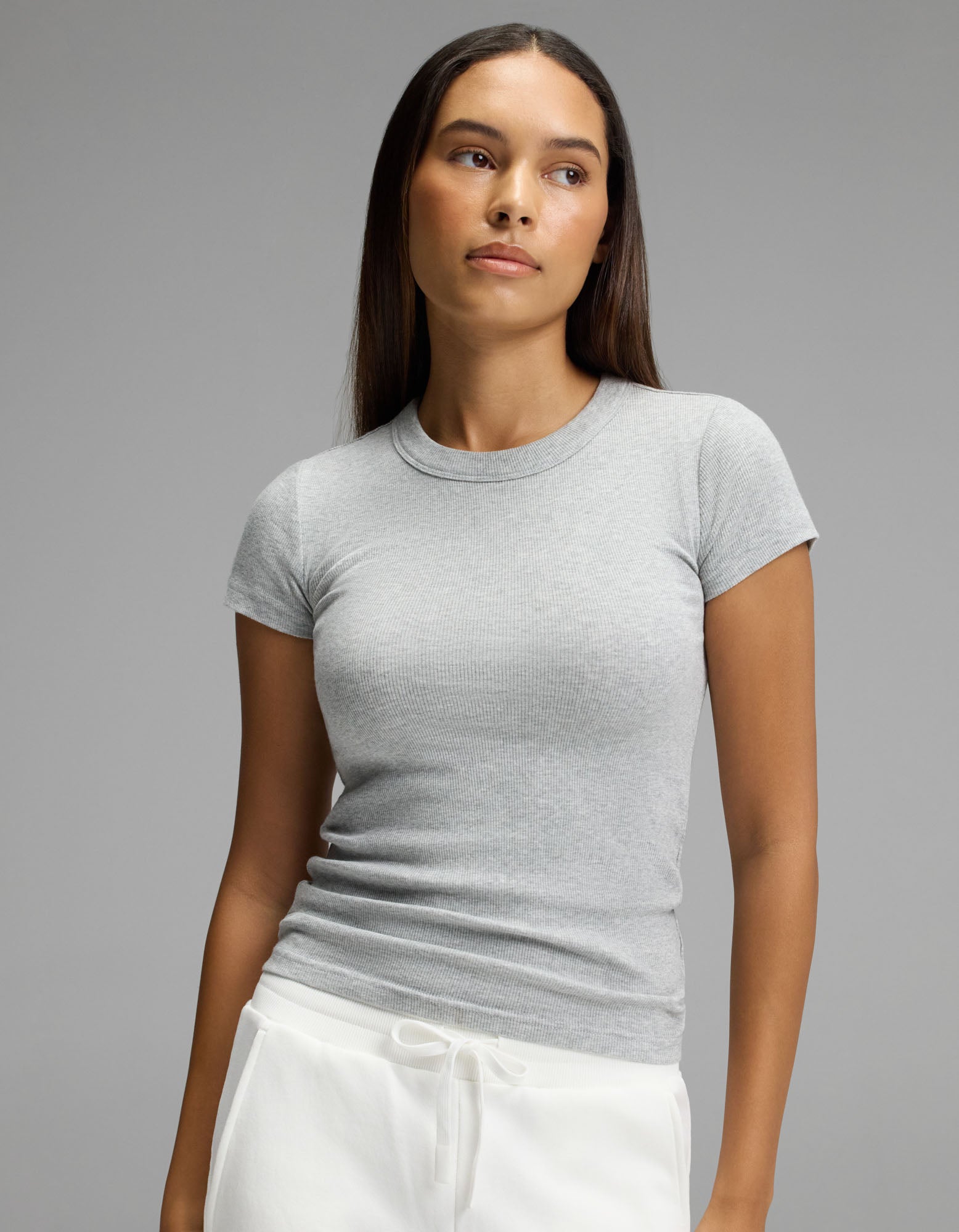 A woman with long brown hair wears the Cozy Earth Women’s Fine Ribbed Tee in light grey and white drawstring pants, standing against a plain grey background and looking slightly to her left with a neutral expression.|Color:Heather Grey