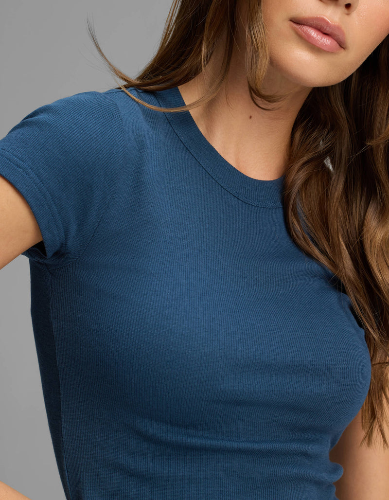 A woman with long brown hair is shown from the torso up, modeling the Cozy Earth Women's Fine Ribbed Tee—a fitted, short-sleeve, blue ribbed t-shirt—against a plain gray background.