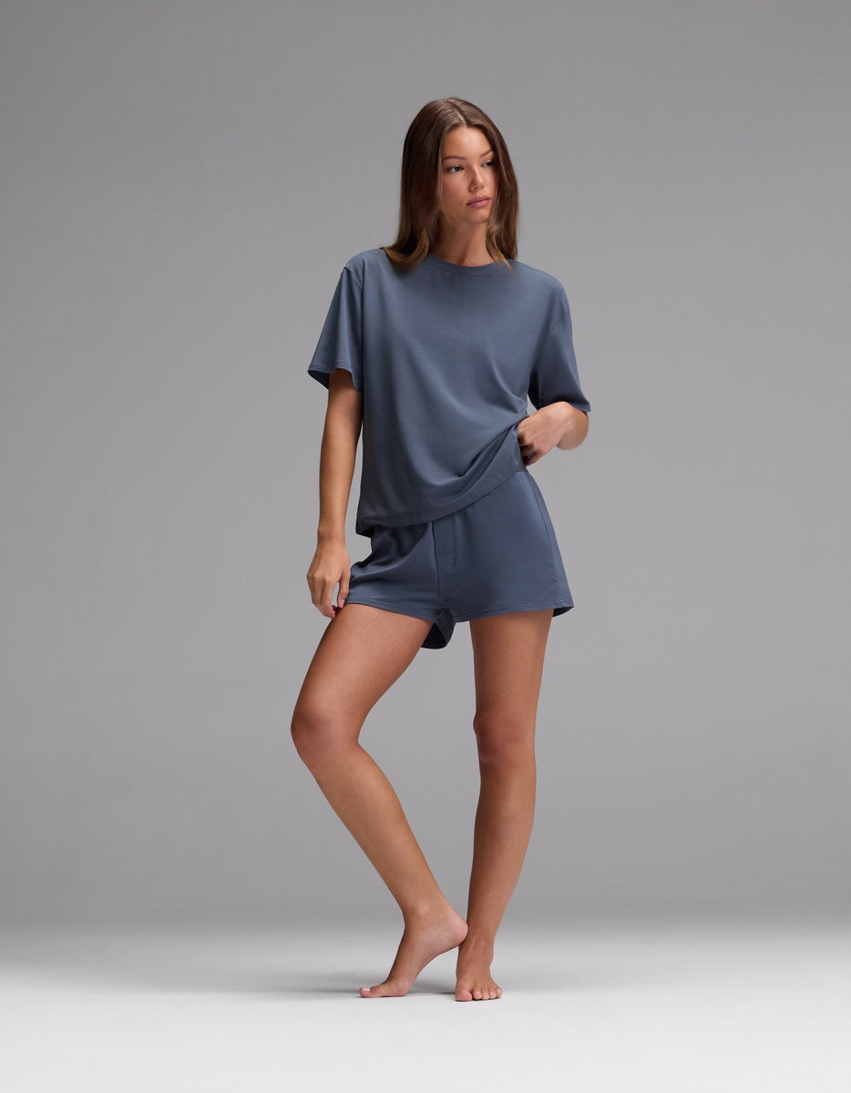 A woman stands barefoot on a gray background, wearing Cozy Earth’s Women’s First Layer Pajama Boxer Short and a matching blue-gray, loose-fitting, short-sleeved top, with one hand in her pocket and the other resting on her thigh.